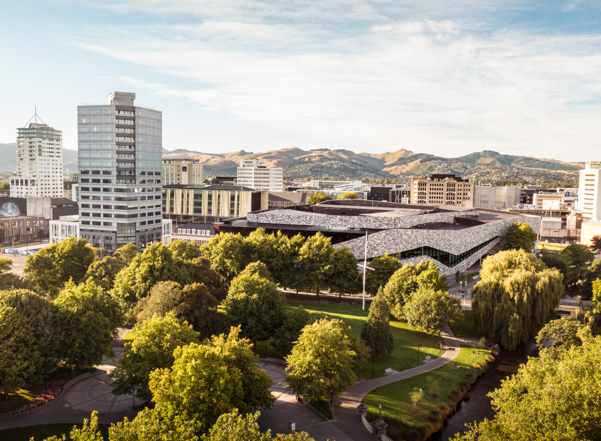 Aerial view of the modern Crowne Plaza Christchurch hotel, towering above a city park, with cityscape and hills beyond.