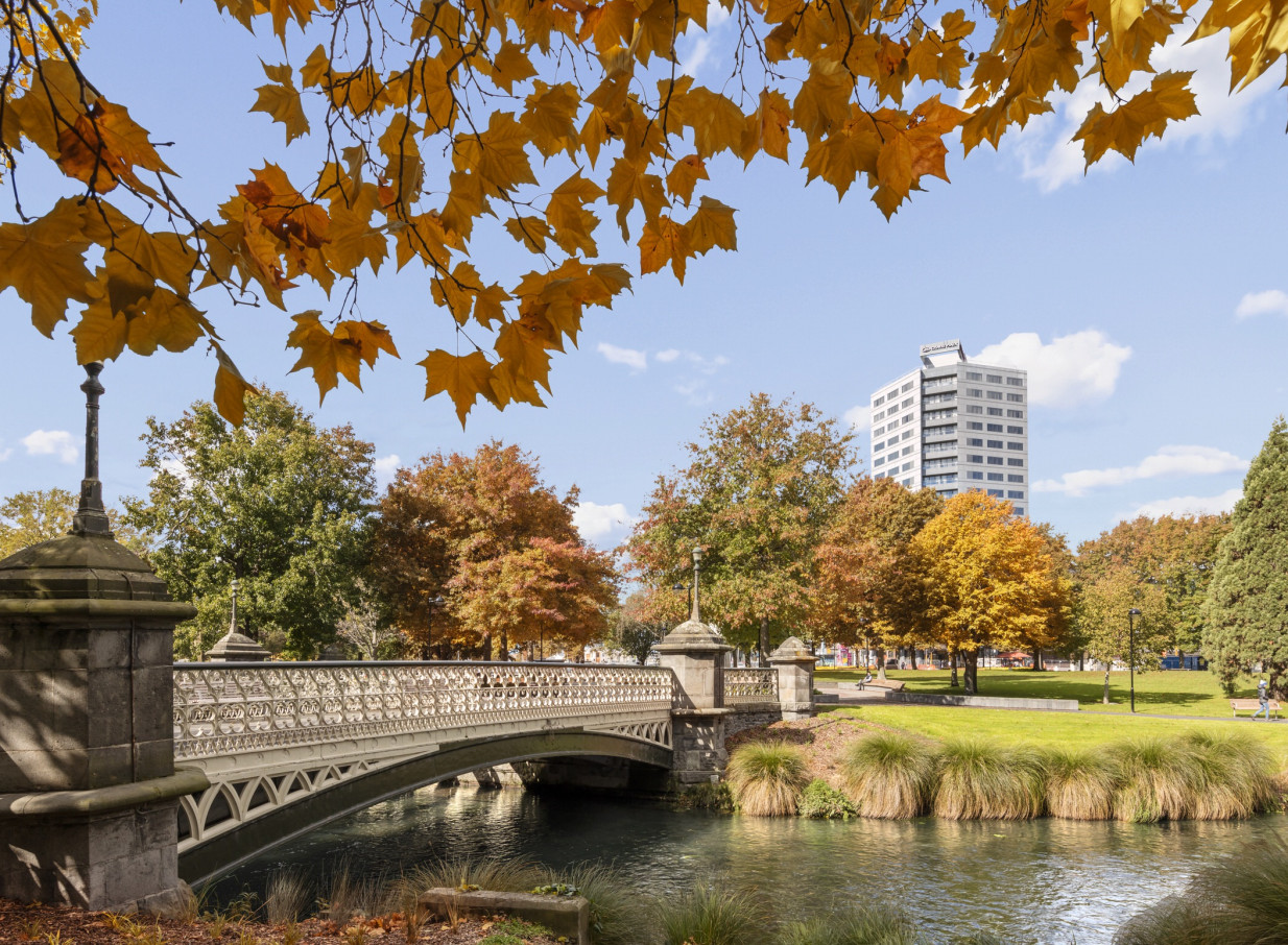 The modern Crowne Plaza Christchurch overlooks a scenic park with an ornate bridge over a river and autumnal trees.