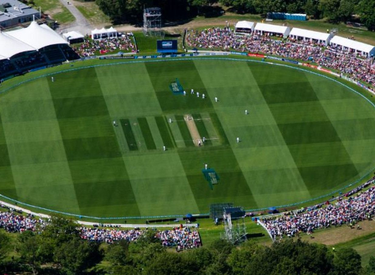 An aerial view captures a cricket match at Hagley Oval in Christchurch, featuring the modern, tent-roofed Hadlee Pavilion.