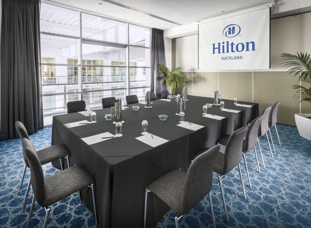 This contemporary boutique-style meeting room at Hilton Auckland features a boardroom setup, a projection screen, and bright windows.