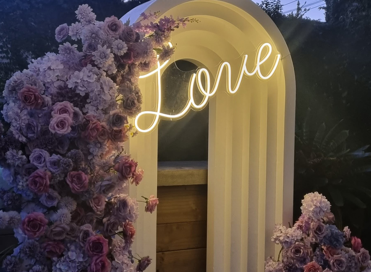 Moxie's elegant Auckland villa offers a charming event arch adorned with a 'Love' neon sign and lush flowers.