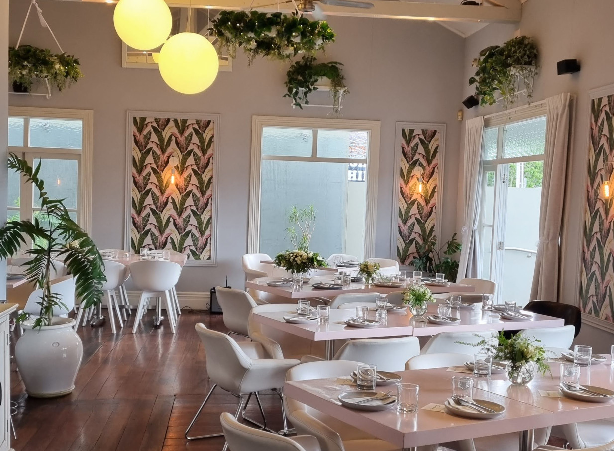 Moxie Auckland's elegant villa dining room features pastel pink tables, white chairs, and exposed rafters, adorned with lush hanging plants.