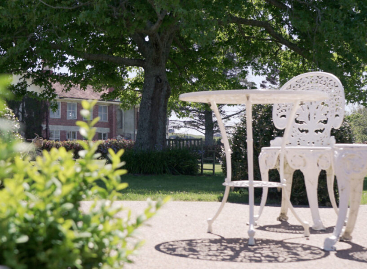 Elegant white wrought-iron furniture sits in the picturesque garden of LaValla Estate, Auckland, with its historic brick building visible.