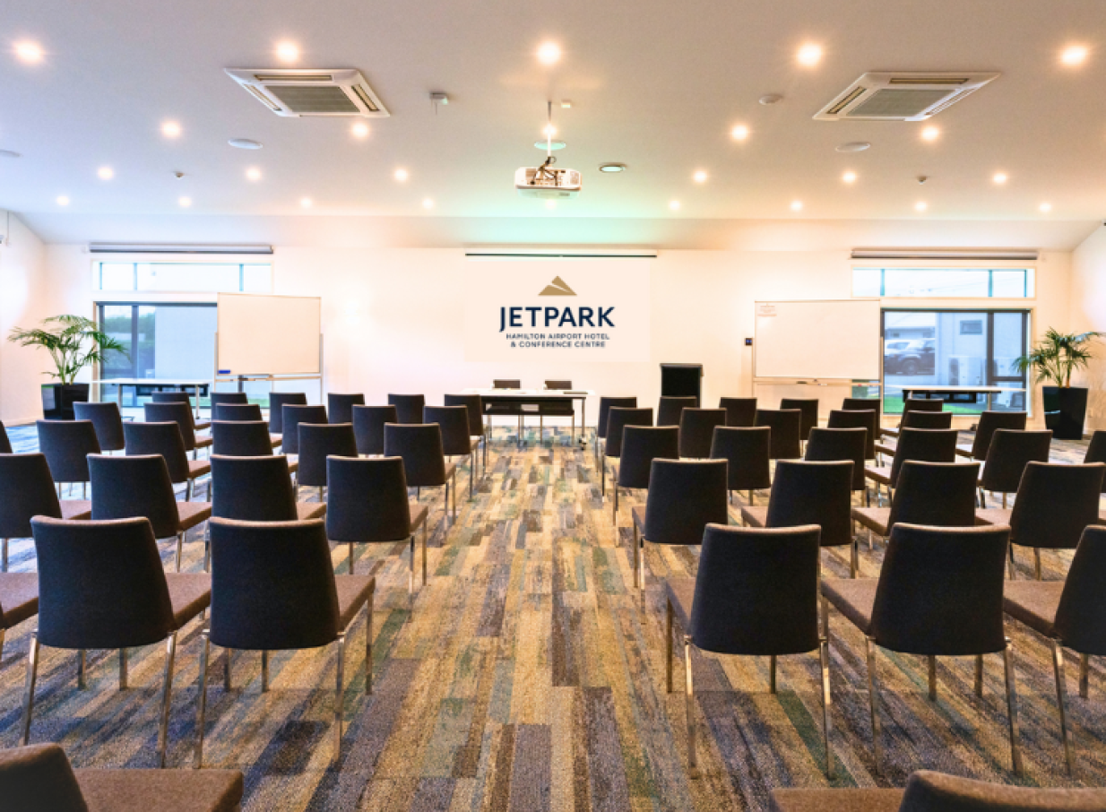 A modern, light-filled conference room with rows of chairs and a projector screen at JetPark Hamilton Hotel.