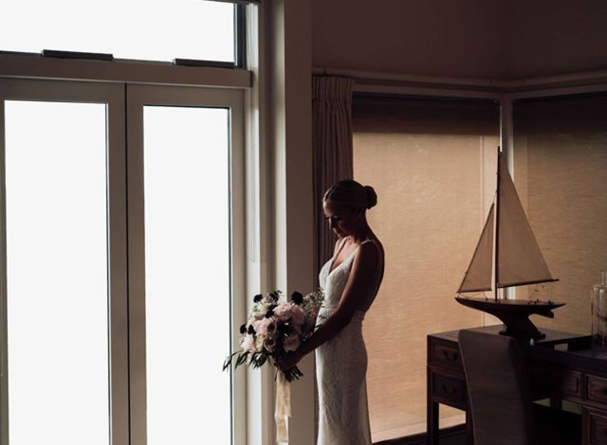 A bride with a bouquet stands in the elegantly styled, light-filled interior of Tu Ngutu Villa in Auckland.