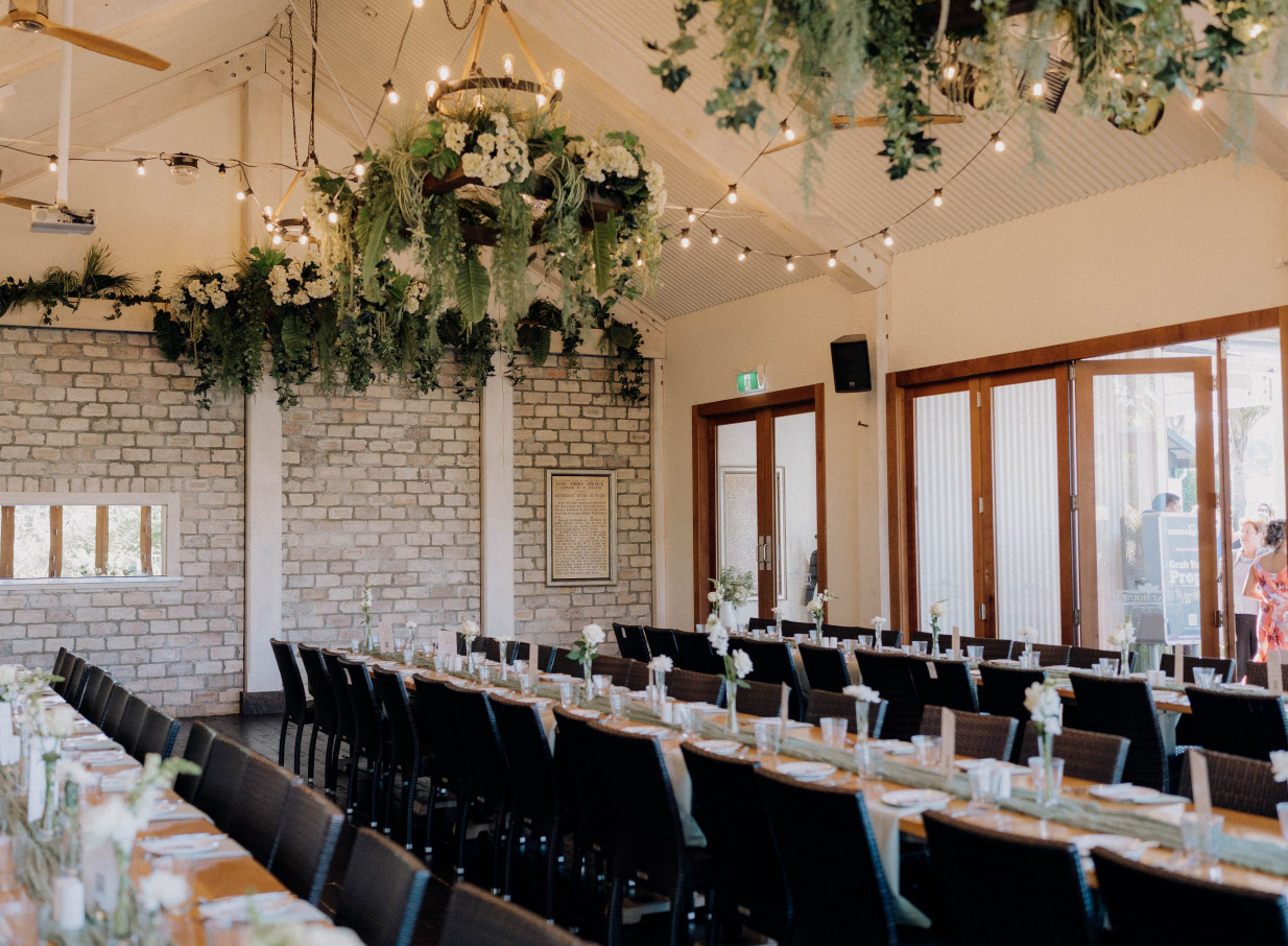 The Boat House in Auckland presents a rustic elegant reception space with long dining tables and lush hanging greenery.