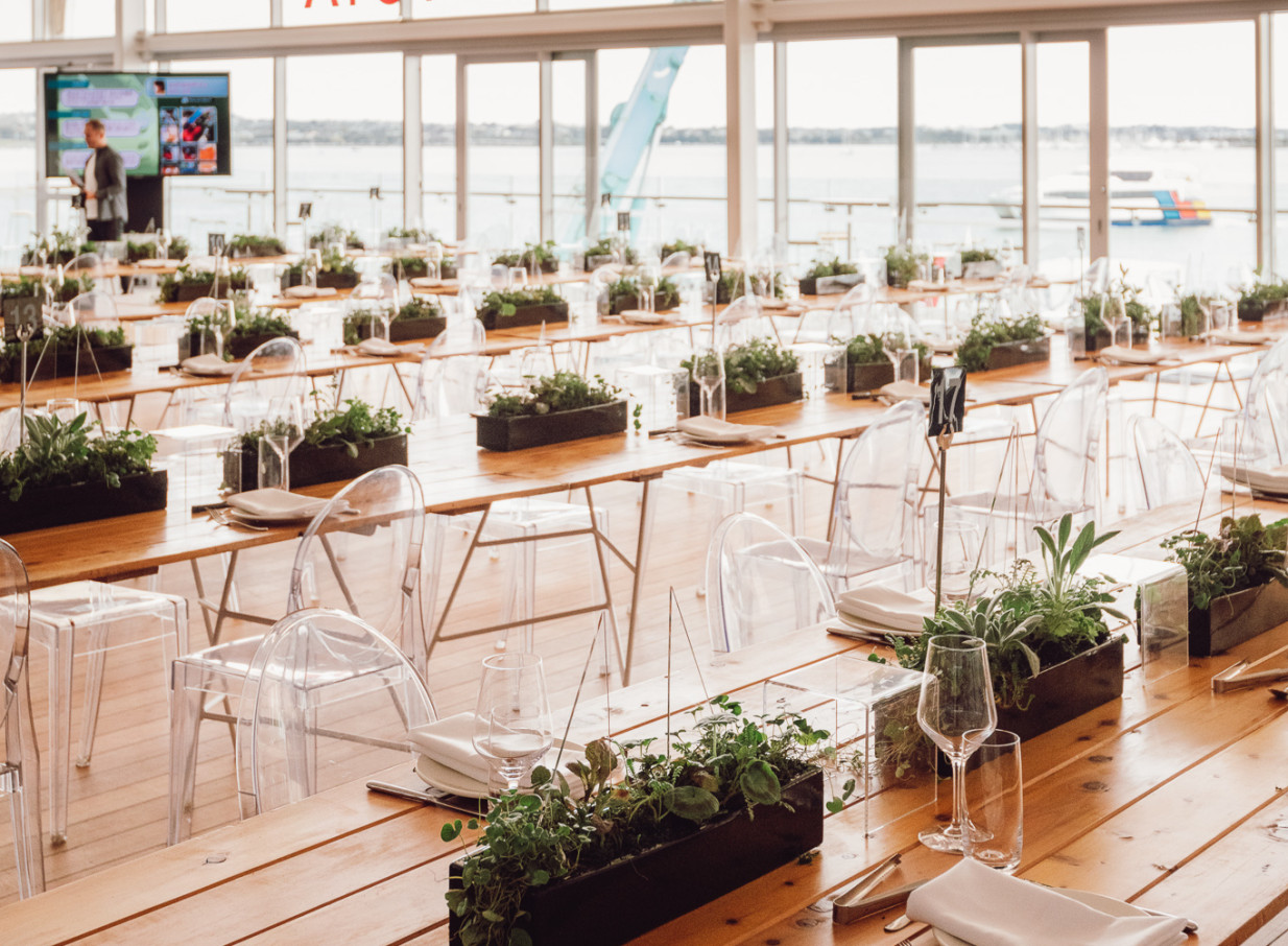 This modern and unique event dining setup at The Cloud, Auckland, features long wooden tables, clear chairs, and harbor views.