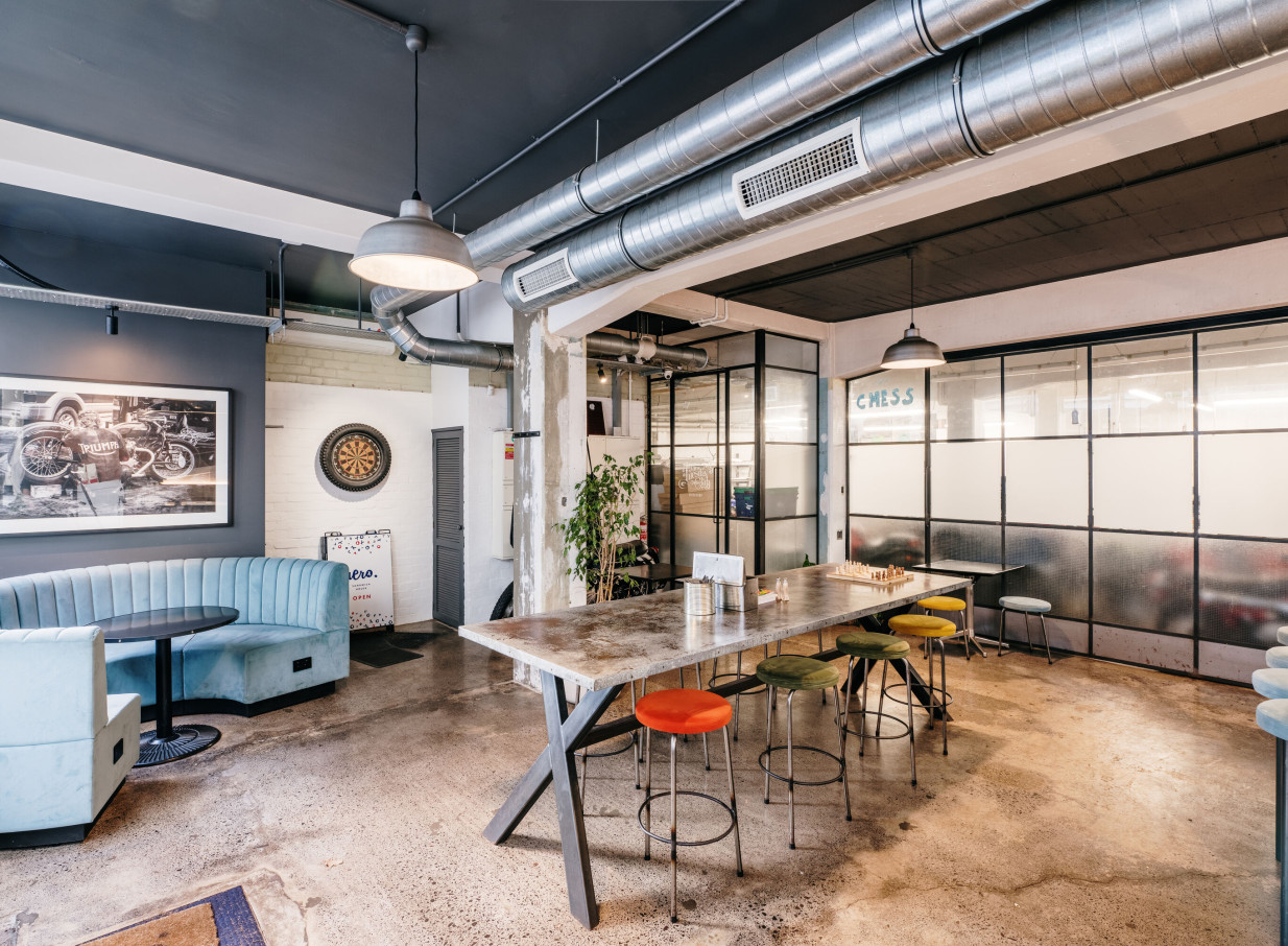 The industrial-chic communal space at The Tuesday Club, Auckland, with exposed pipes, concrete floors, and varied seating.