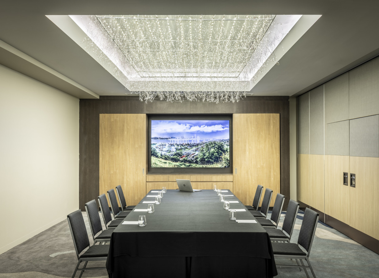An elegant boardroom at Grand Millennium Auckland, featuring a long conference table, large screen, and a modern, sparkling light fixture.