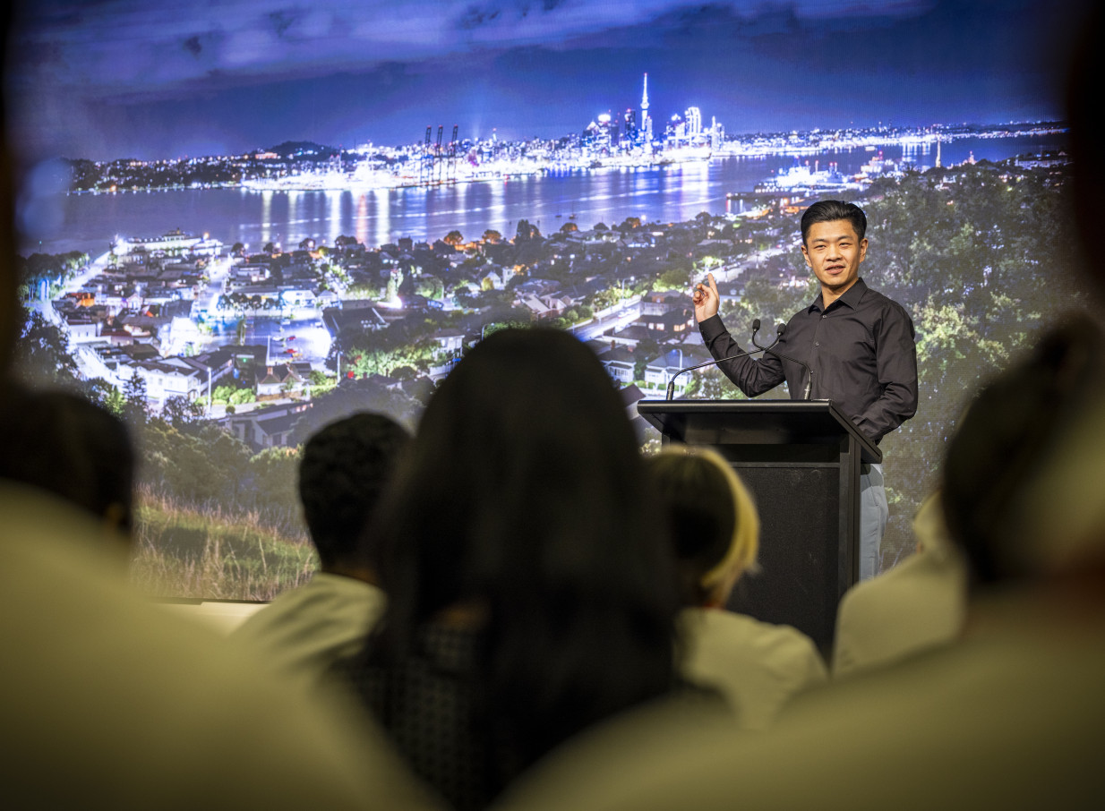A speaker presents in an elegant, modern conference space at Grand Millennium Auckland, featuring Auckland's illuminated city skyline on a large screen.