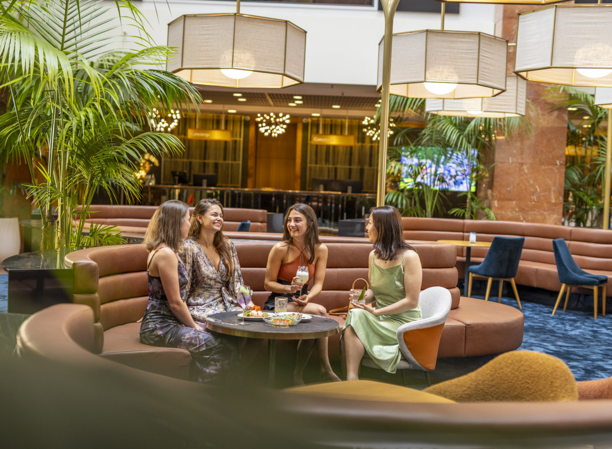 Friends enjoy drinks in the chic lobby lounge of Grand Millennium Auckland, featuring modern seating and lush plants.