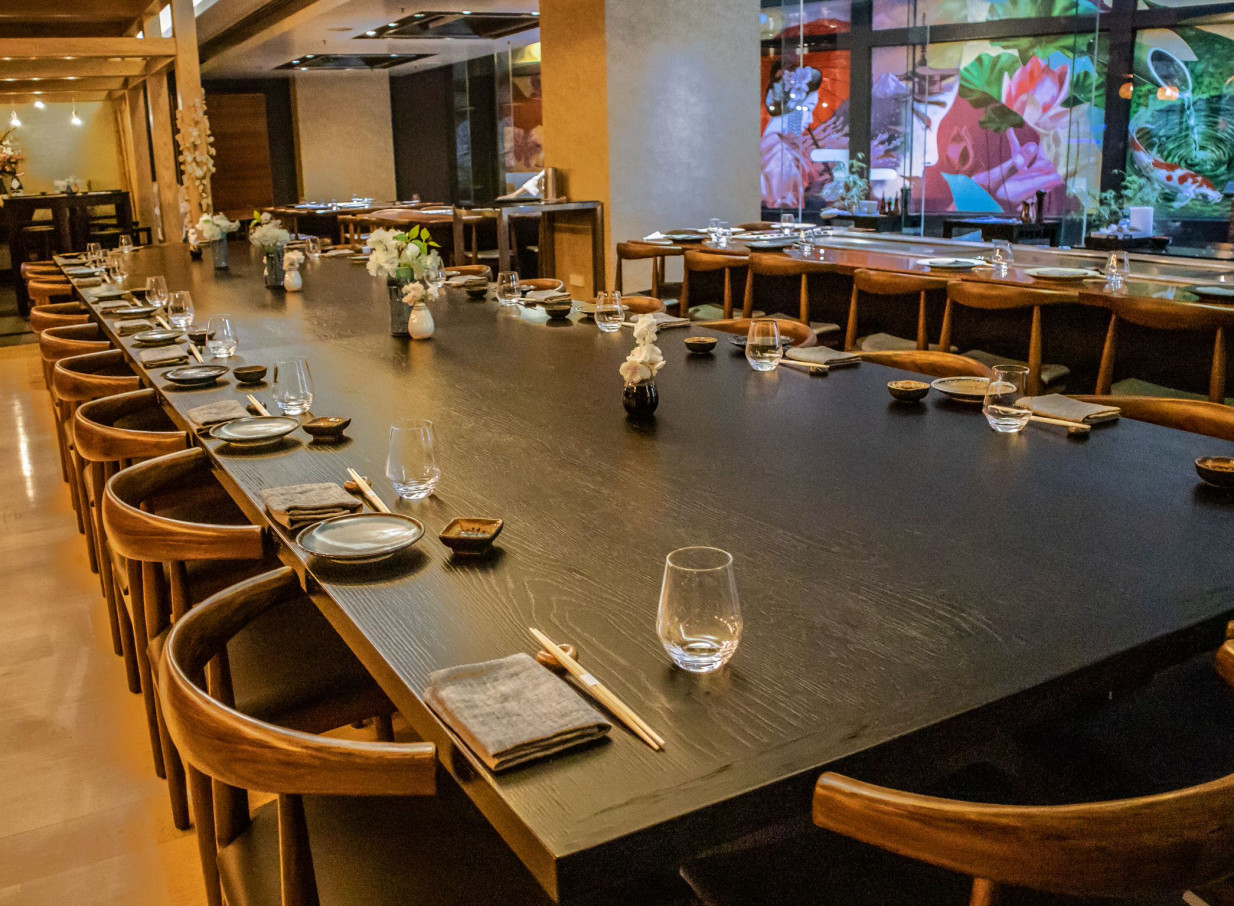 A contemporary Japanese dining room at Grand Millennium Auckland features a long dark communal table with curved wooden chairs and vibrant background art.