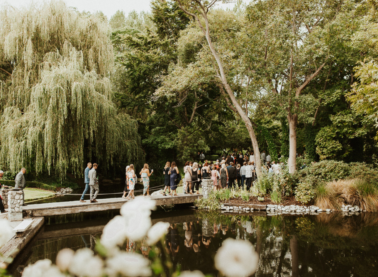 Guests cross a wooden bridge over a tranquil pond at Stonebridge, a natural garden wedding venue in South Canterbury.