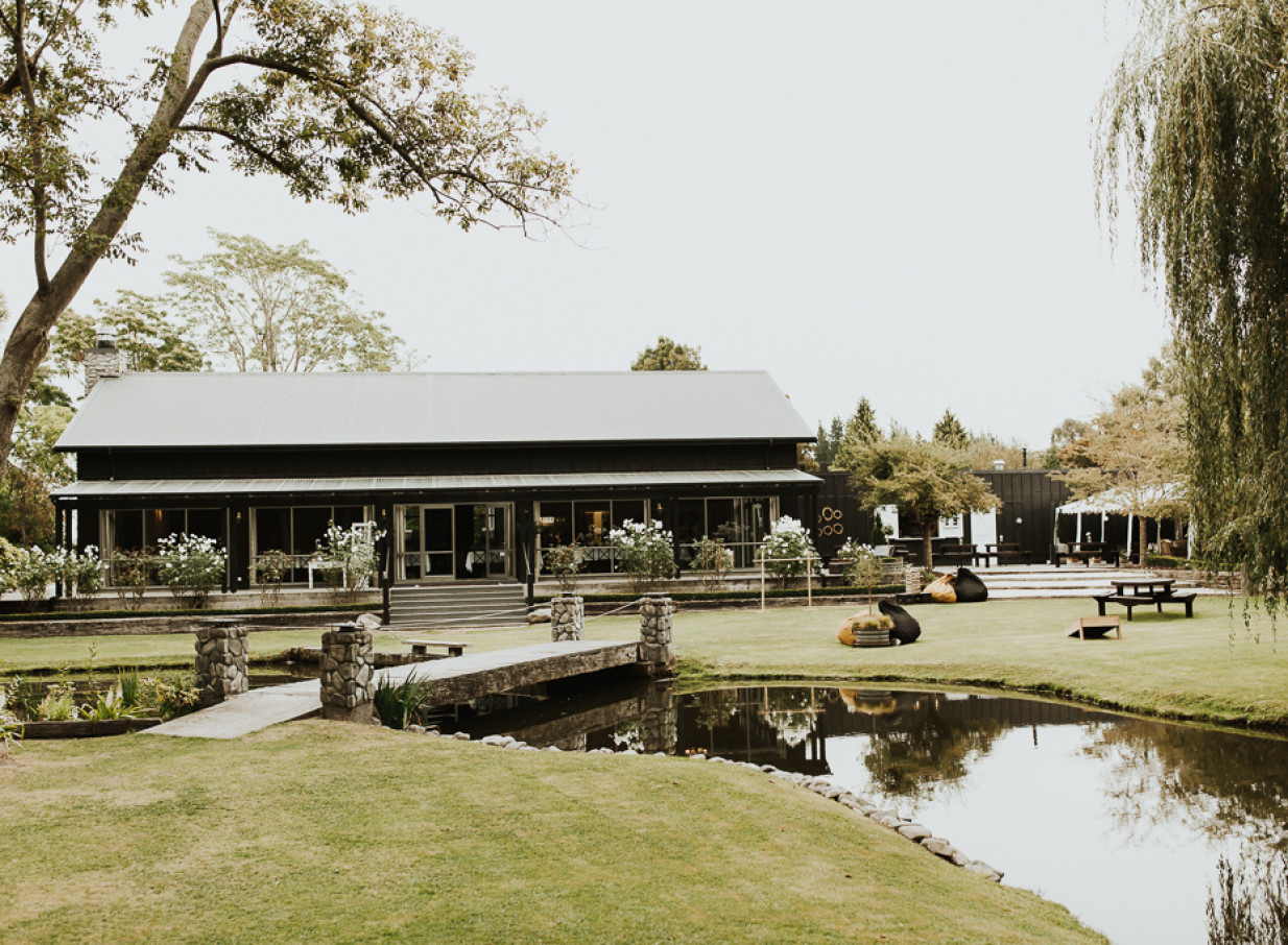 The modern event lodge at Stonebridge, Canterbury, is nestled among manicured gardens, featuring a tranquil pond and stone bridge.
