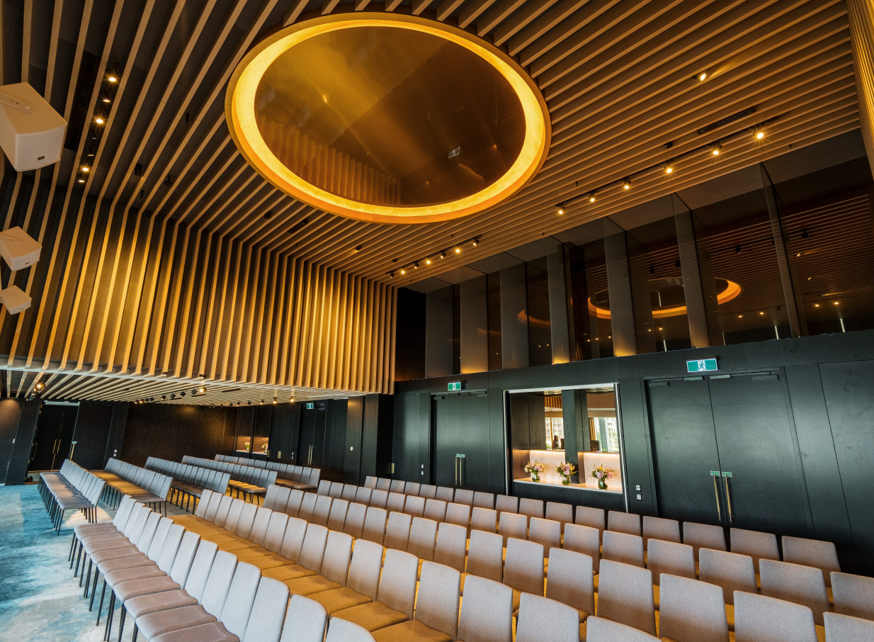 An elegant event hall at Park Hyatt Auckland, featuring a modern slatted timber ceiling with a prominent golden circular light, arranged for a presentation.