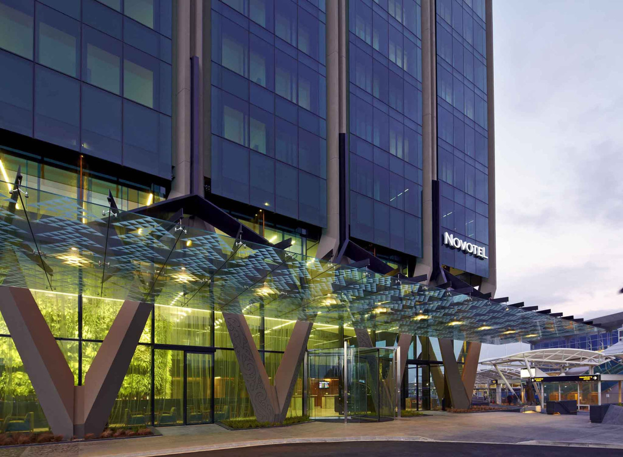 The modern Novotel Auckland Airport entrance features a fritted glass canopy, V-columns reflecting Maori design, and an interior green wall.