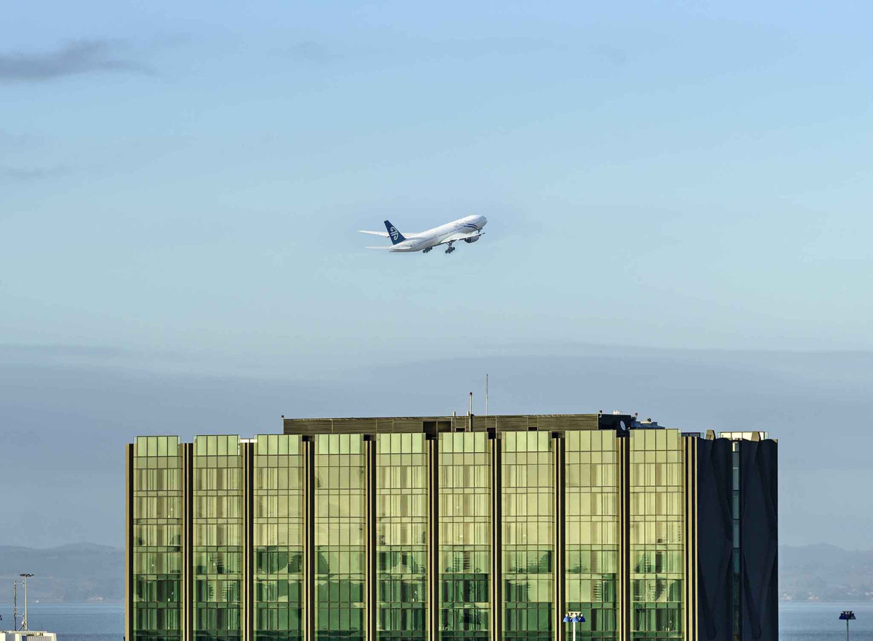 An airplane takes off over the Novotel Auckland Airport, a modern building featuring a reflective green glass facade.