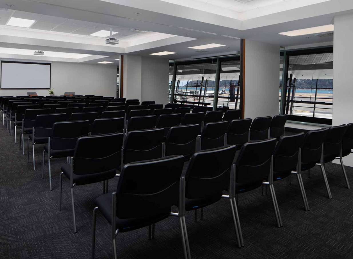A modern, theatre-style conference room at Rydges Wellington Airport, featuring large windows with views of the airport terminal and runway.