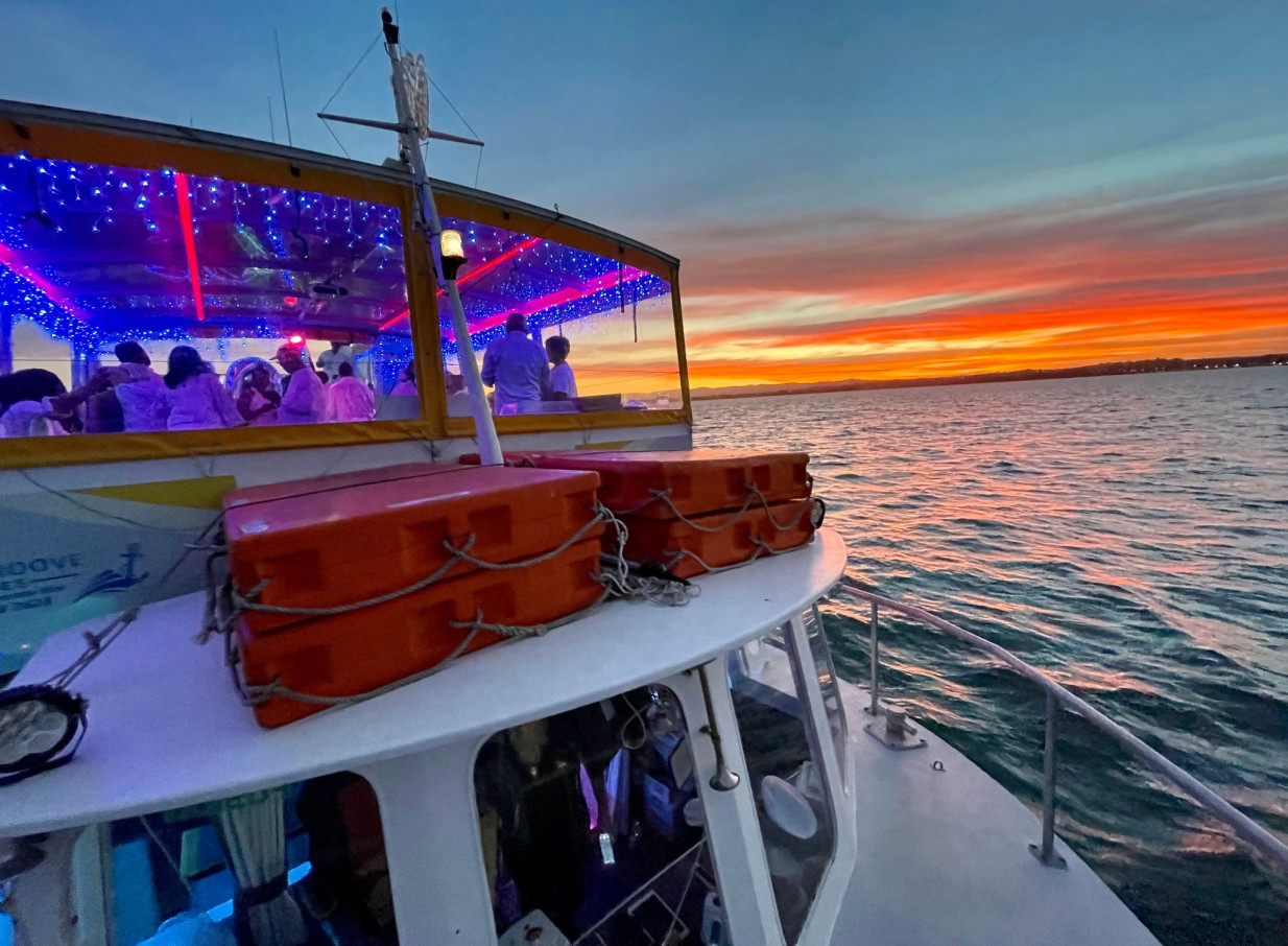 The vibrant party cruise interior of Ocean Groove Cruises in Auckland is illuminated by blue lights, set against a stunning sunset over the water.