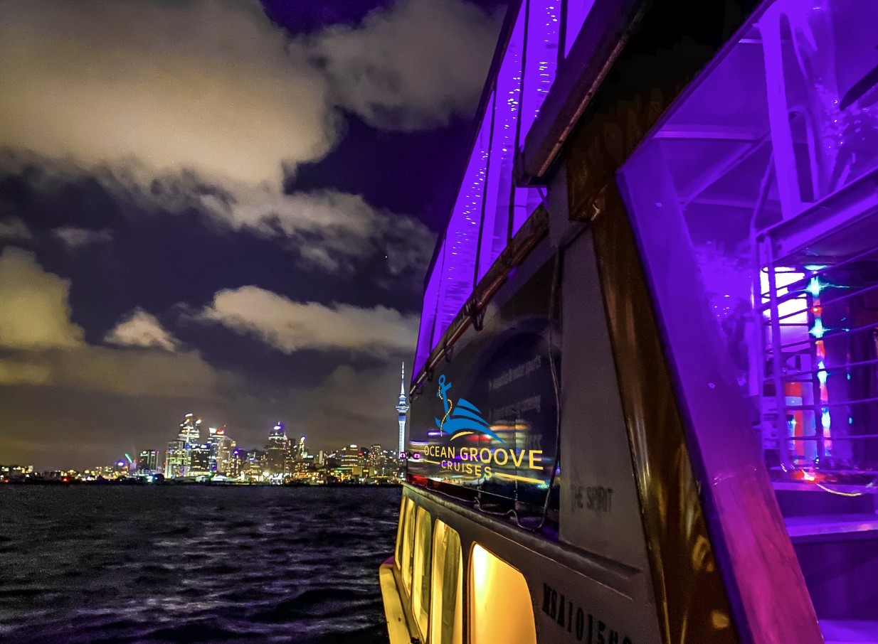 Ocean Groove Cruises' purple-lit party boat cruises Auckland Harbour against the sparkling city skyline.