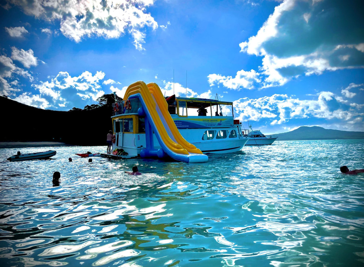 Guests enjoy a large inflatable slide off an Ocean Groove Cruises party boat in Auckland's clear waters.