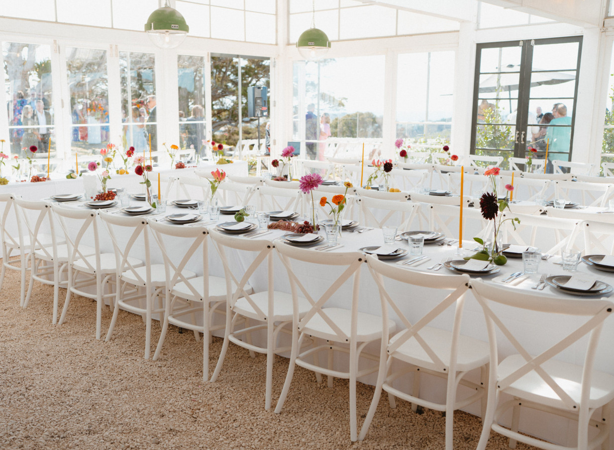 The light-filled Greenhouse & Terrace event space at Batch Winery, Waiheke Island, is set with white cross-back chairs and vibrant floral centerpieces.