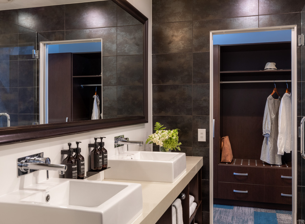 A sleek, modern hotel bathroom at Rydges Auckland, featuring dual vessel sinks, dark tiled walls, and an adjacent open closet.