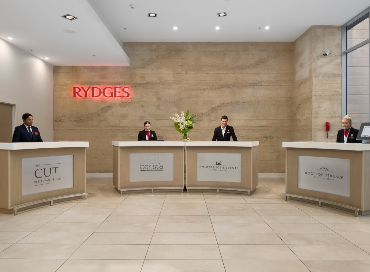 The contemporary hotel lobby at Rydges Auckland features multiple service desks against a textured wall with a glowing red sign.