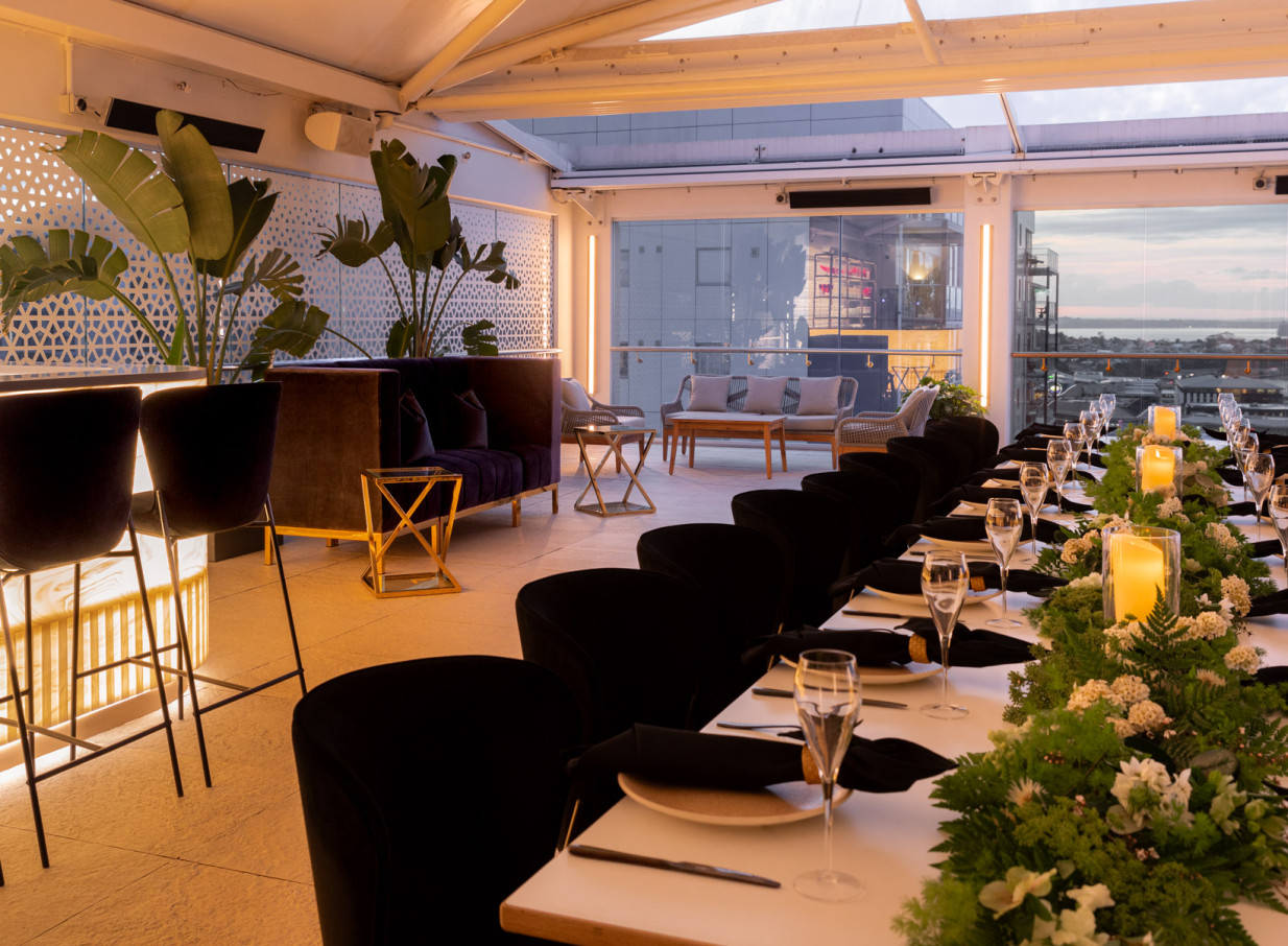 Rydges Auckland's modern rooftop terrace hosts a formal dinner setup and chic lounge areas with panoramic city views.