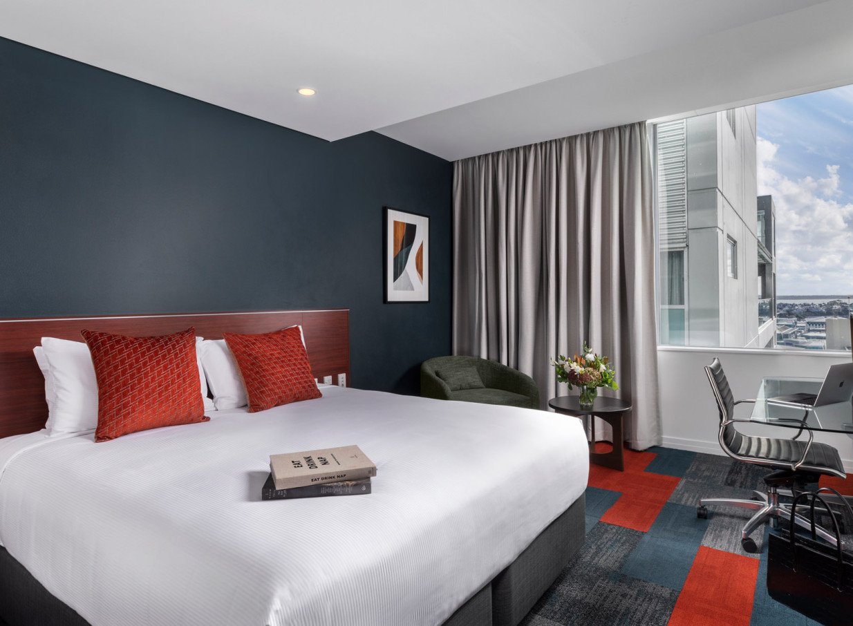 A modern hotel bedroom at Rydges Auckland features a large bed, workspace, and a city view.