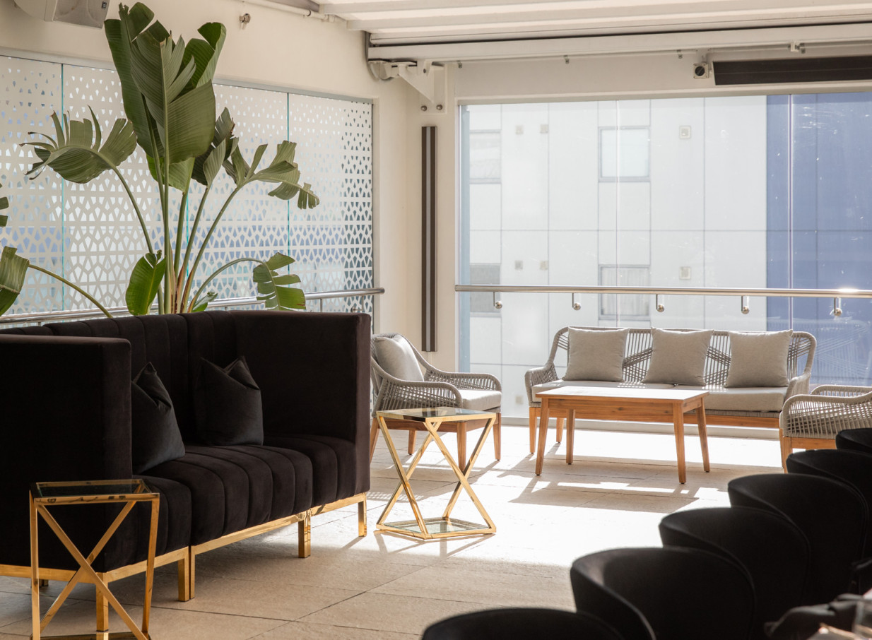 Rydges Auckland's chic rooftop lounge offers a contemporary urban retreat with modern seating, a large plant, and glass walls.