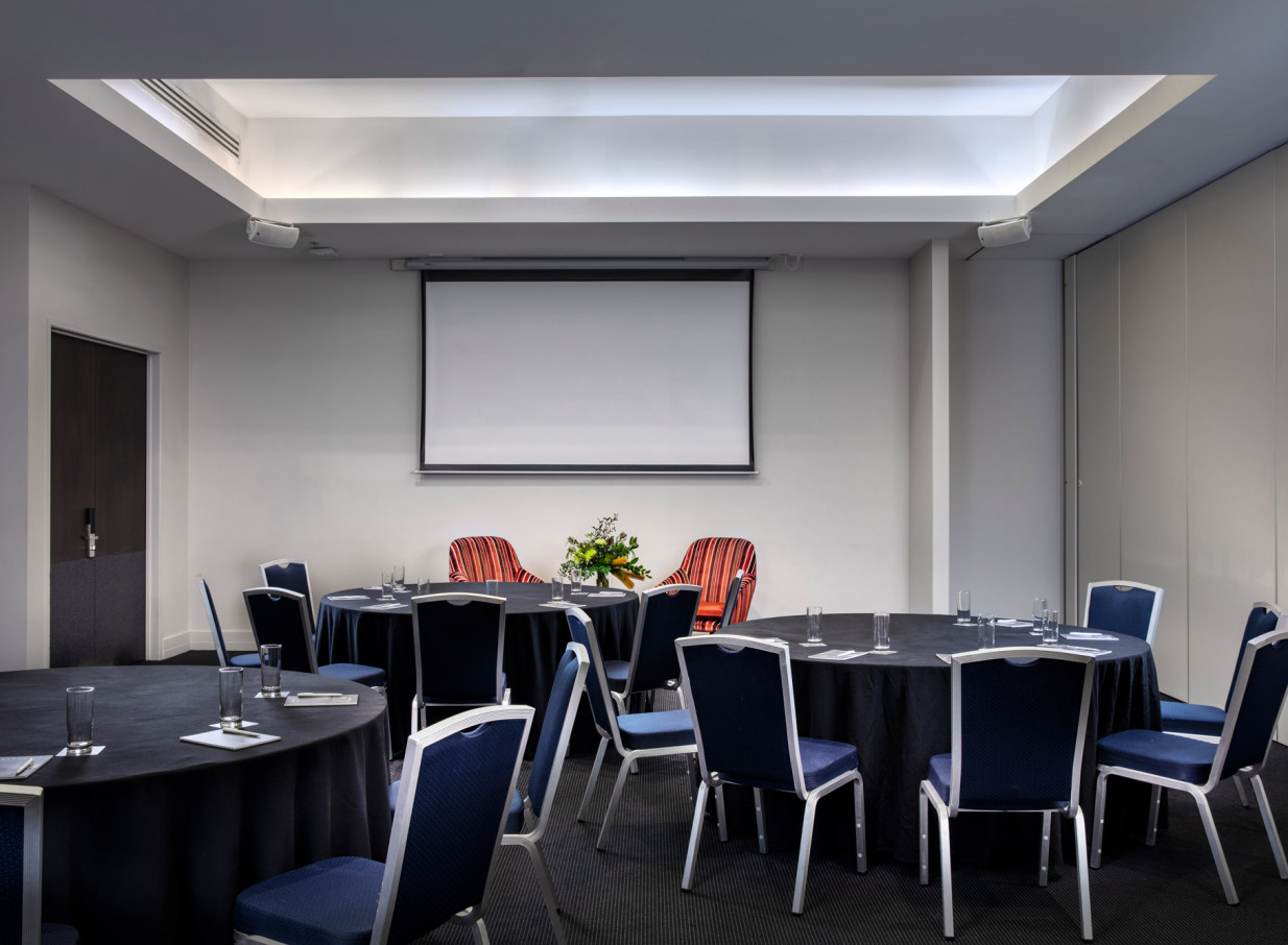 A contemporary meeting room at Rydges Auckland, featuring round tables, blue chairs, and a projector screen for events.