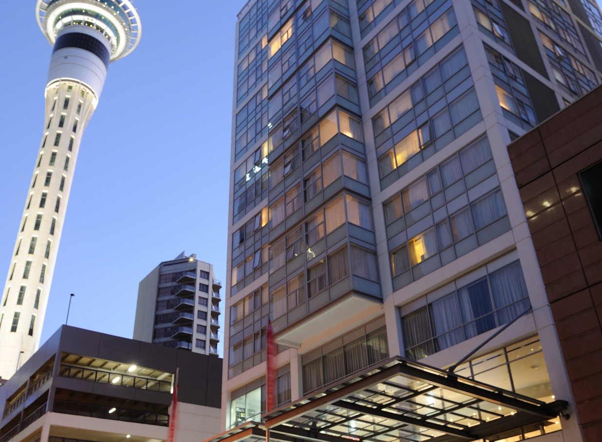 The contemporary Rydges Auckland hotel exterior at dusk, featuring a sleek, glass-fronted design next to the Sky Tower.