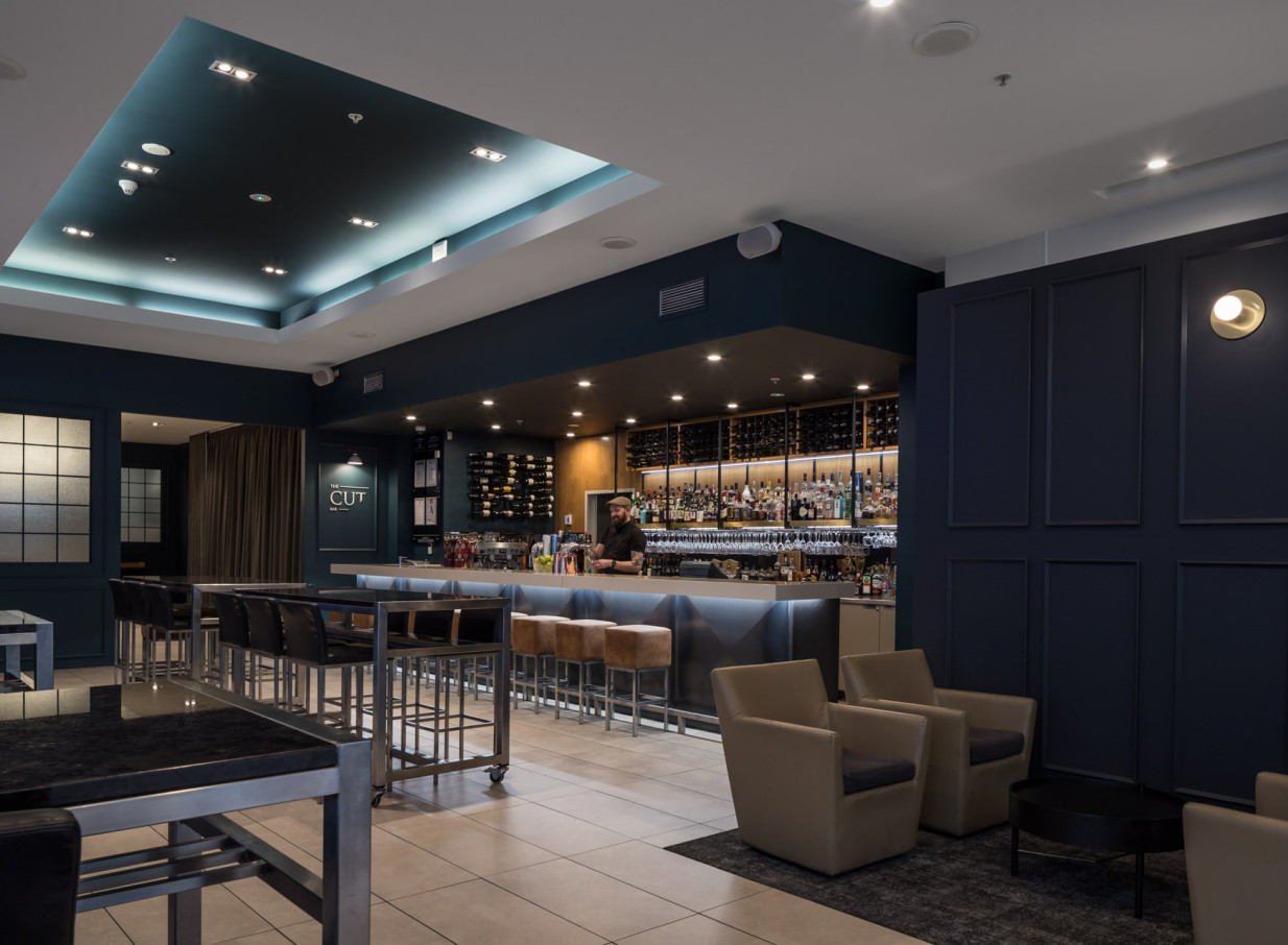 A contemporary bar area at Rydges Auckland's The CUT, featuring a luminous backlit counter, wine display, and lounge seating.