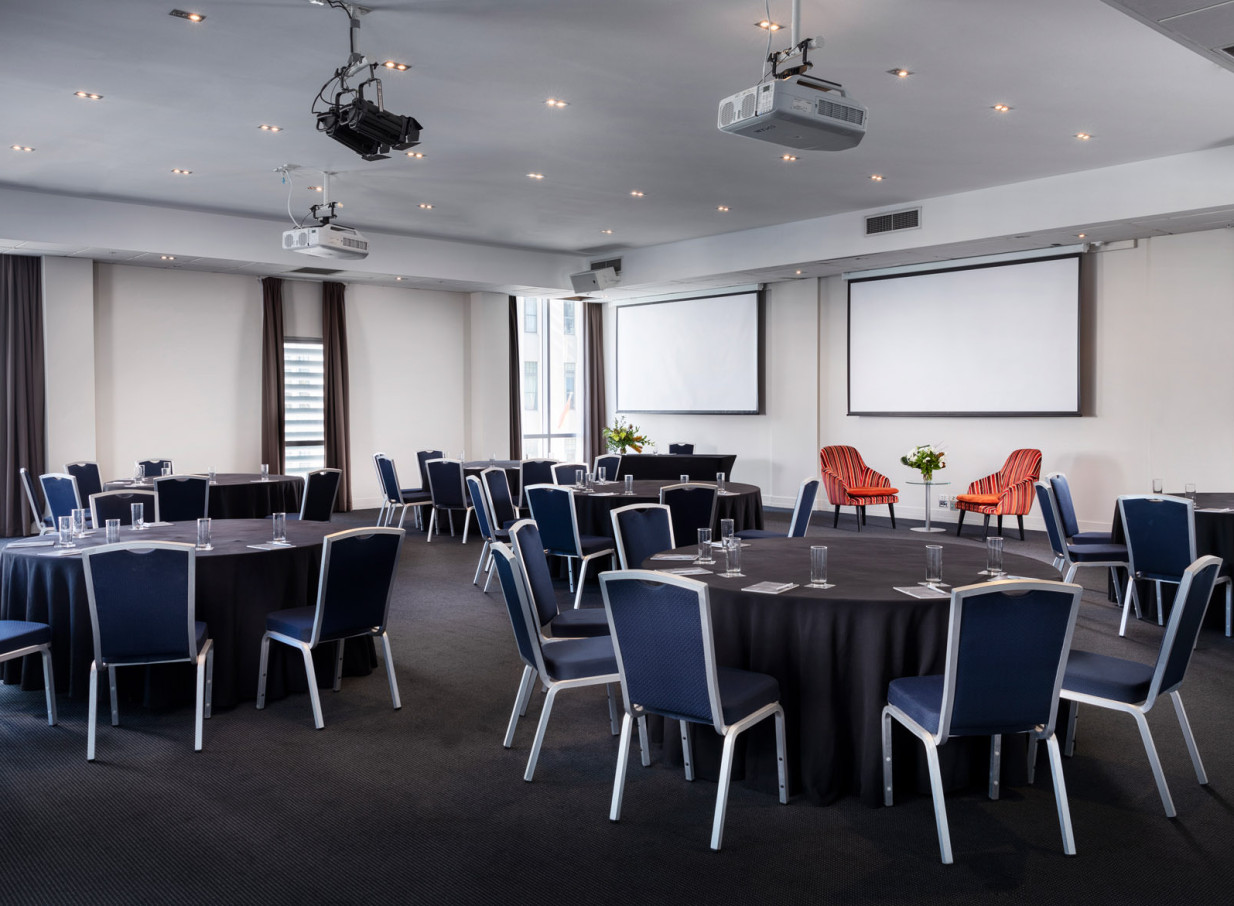 Rydges Auckland's contemporary conference room in Auckland, set with round tables, blue chairs, and dual projection screens.