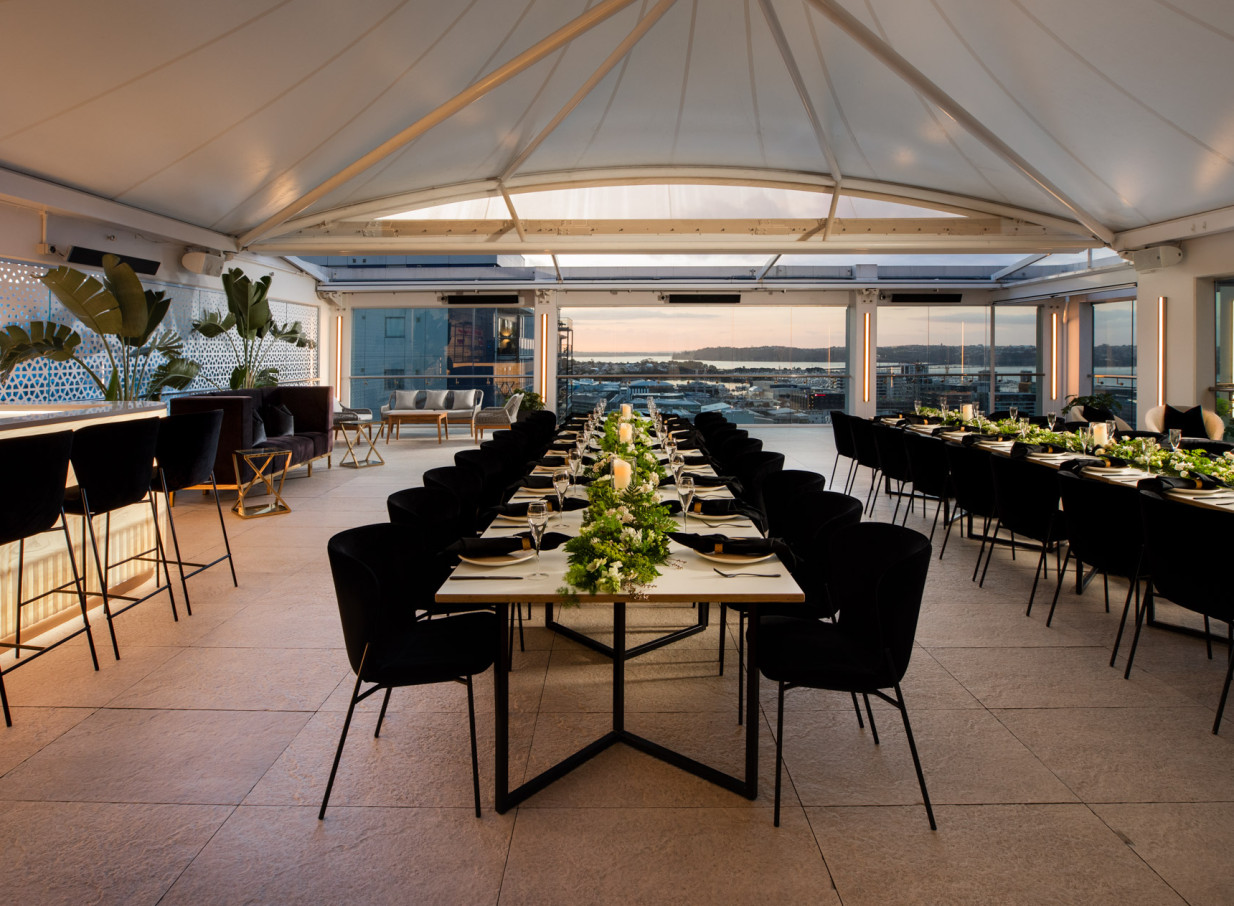 Rydges Auckland's modern rooftop event space showcases a long dining table under a retractable roof with panoramic city views.