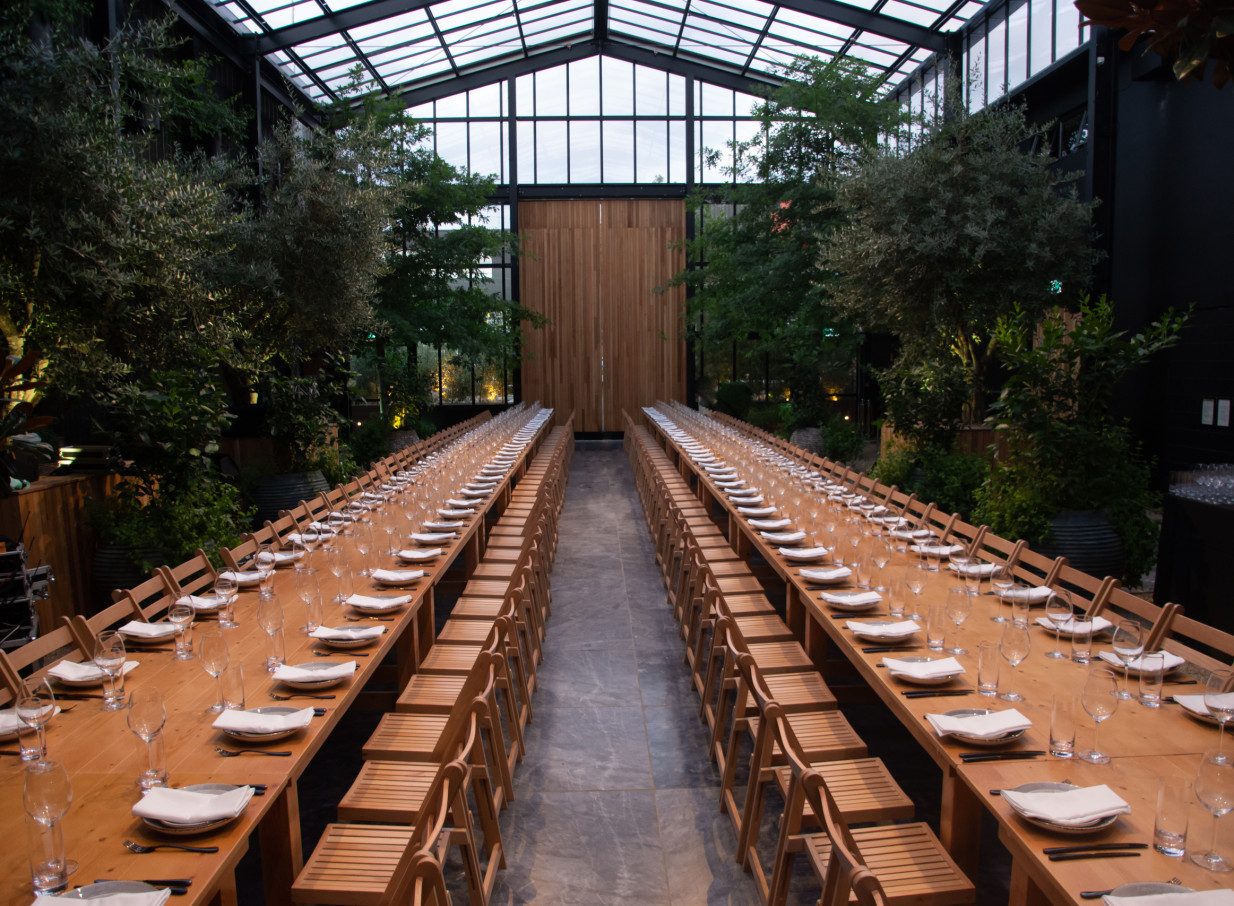 Long banquet tables fill the industrial-chic, glass-walled event space at Glasshouse Morningside, Auckland, surrounded by lush greenery.