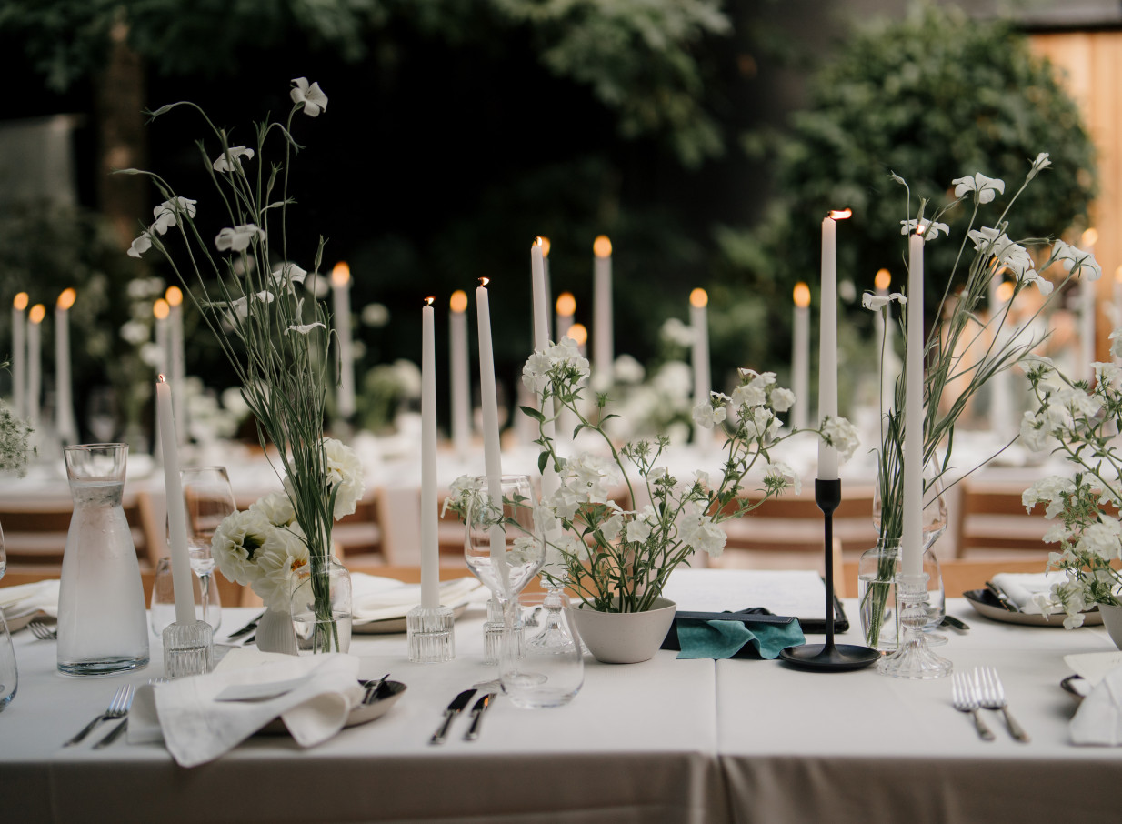 An elegant dining table with candles and white florals is set within the industrial-chic, glass-walled Glasshouse Morningside, Auckland.