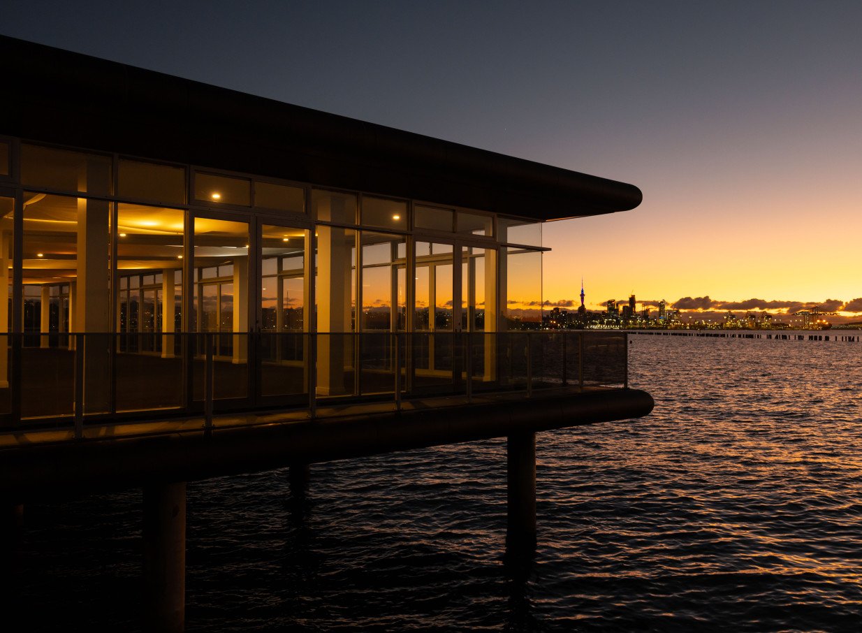 OKAHU in Auckland offers a modern waterfront event space with panoramic glass walls, overlooking the city skyline at sunset.