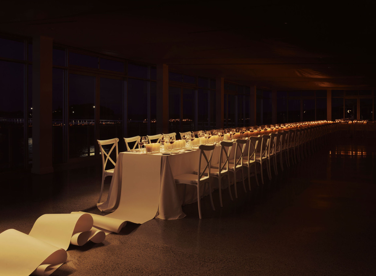 A candlelit long dining table in OKAHU, Auckland's modern waterfront event space, featuring panoramic harbor views.