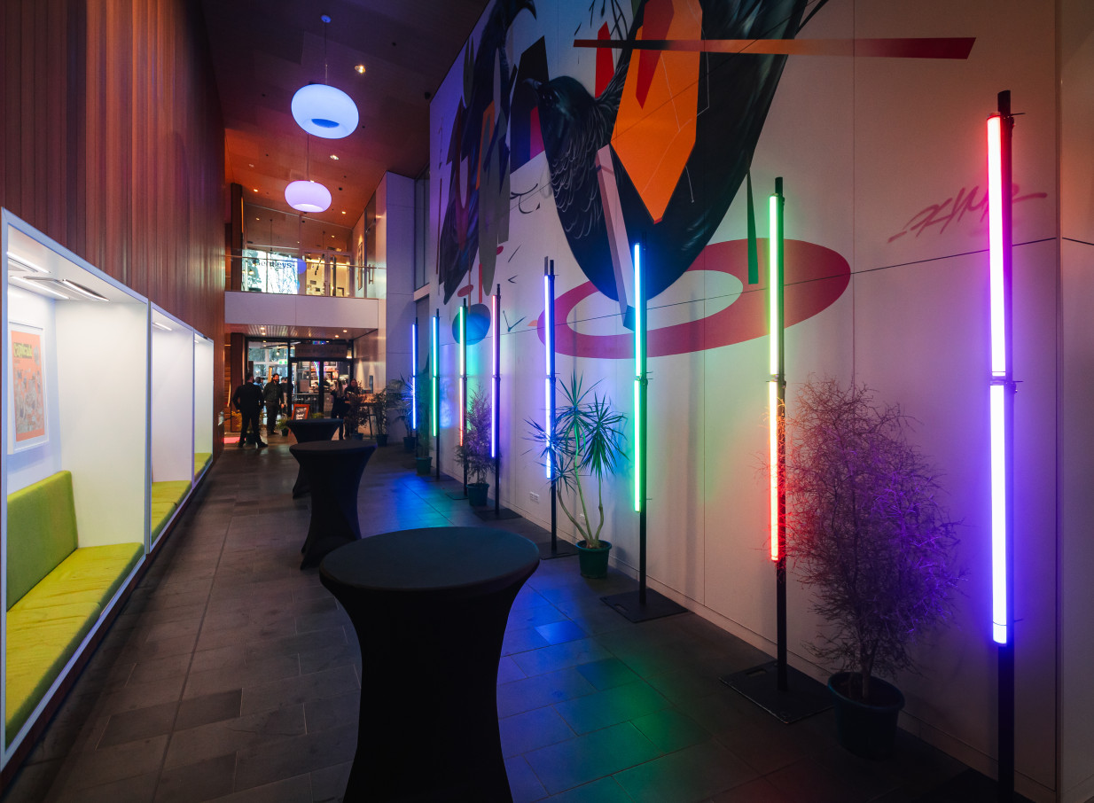 Haere-roa, a modern event space in Christchurch, features a vibrant mural and colorful vertical lights with high-top tables.