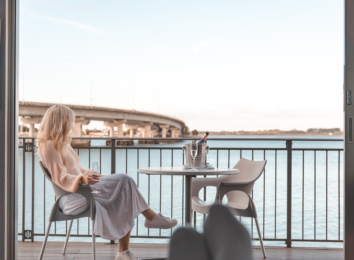 A woman relaxes with champagne on an outdoor balcony with panoramic water views at the chic, over-water Trinity Wharf hotel in Tauranga.