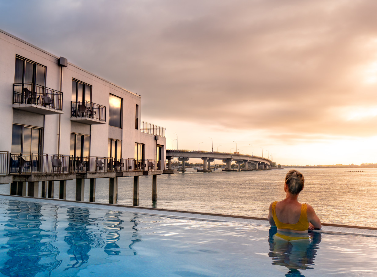 A person enjoys the infinity pool at Trinity Wharf Tauranga, a modern over-water hotel, overlooking the harbour at sunset.