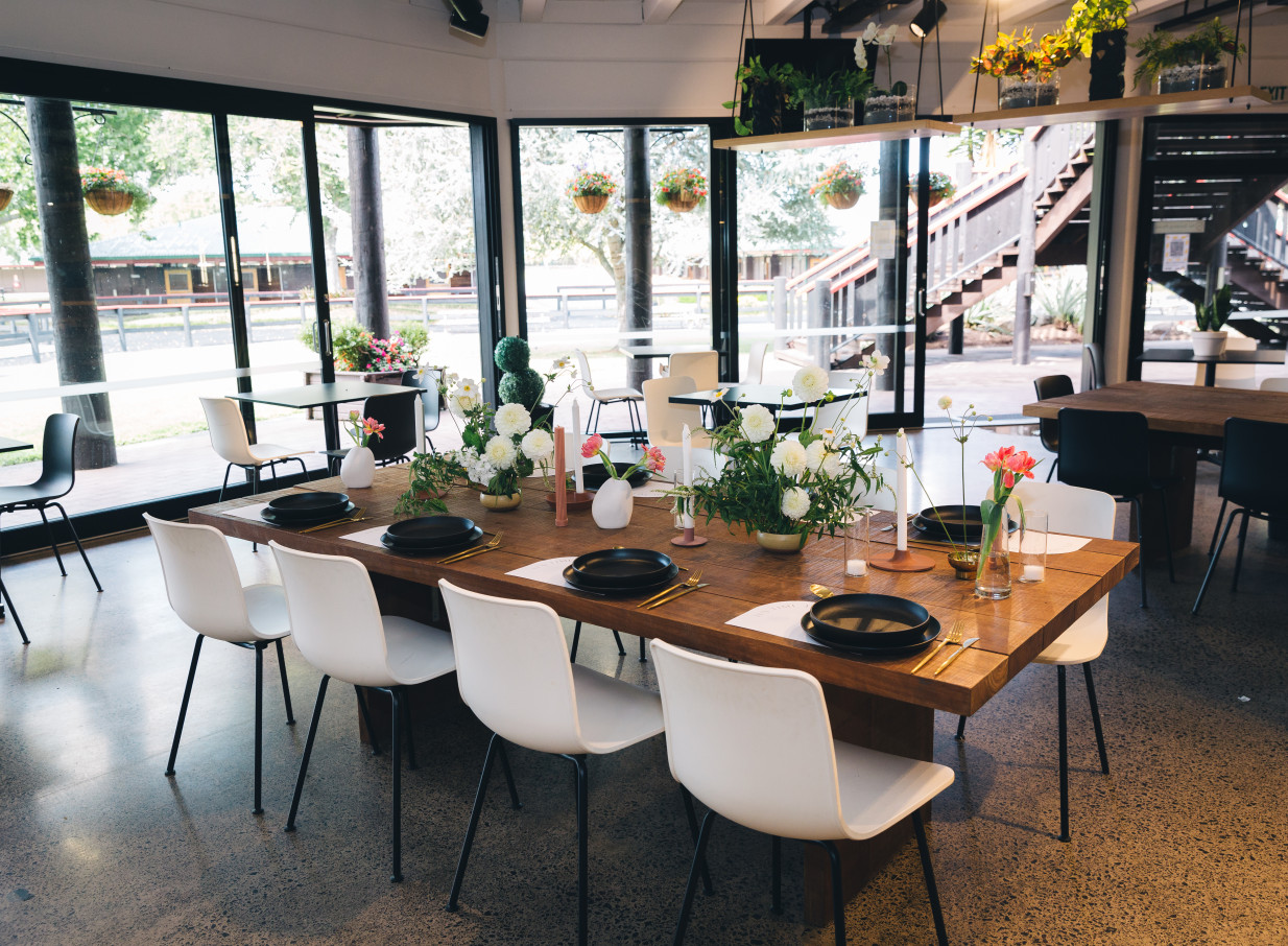 An airy, modern dining area at Karaka Pavilion in Auckland, with a long wooden table set amidst views of lush gardens.