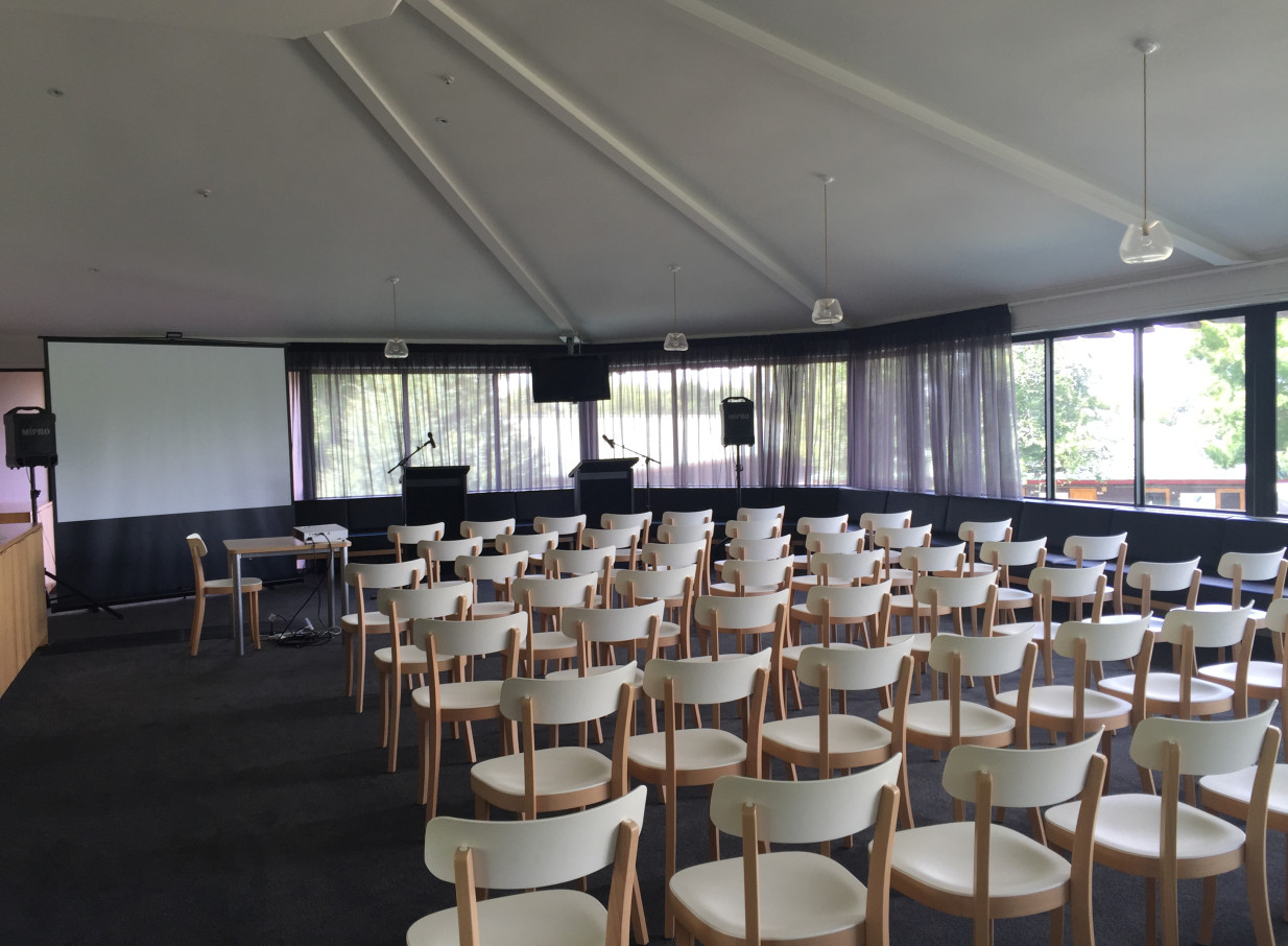 This modern conference setup, featuring white chairs and a large screen, is found within the high-ceilinged Karaka Pavilion in Auckland.