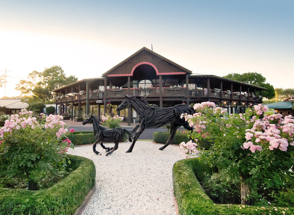 The image displays the exterior of the Karaka Pavilion in Auckland, an equestrian-themed event venue, featuring its grand wooden architecture, horse sculptures, and manicured rose gardens.