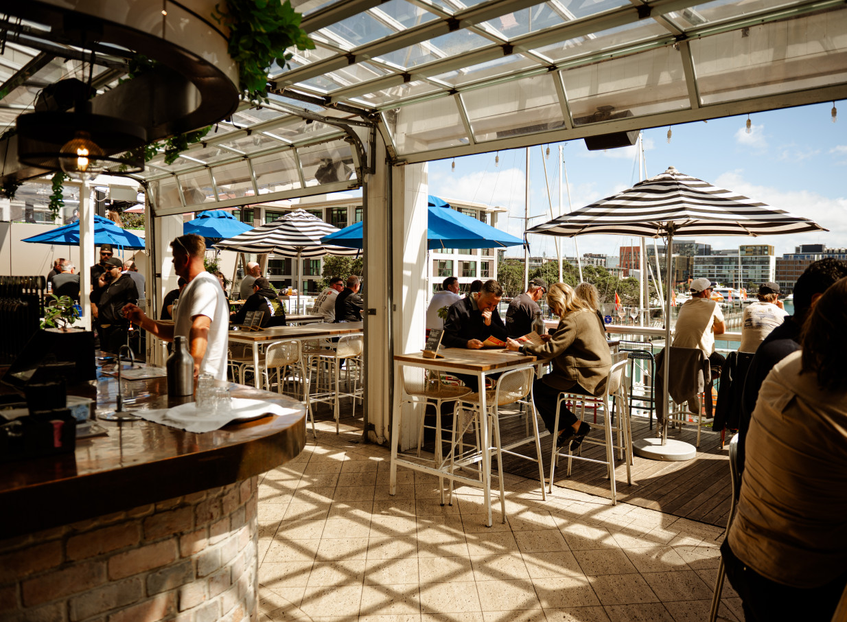 Patrons enjoy the vibrant, open-air rooftop bar at Dr Rudi's in Auckland, offering clear views of the Viaduct Harbour.