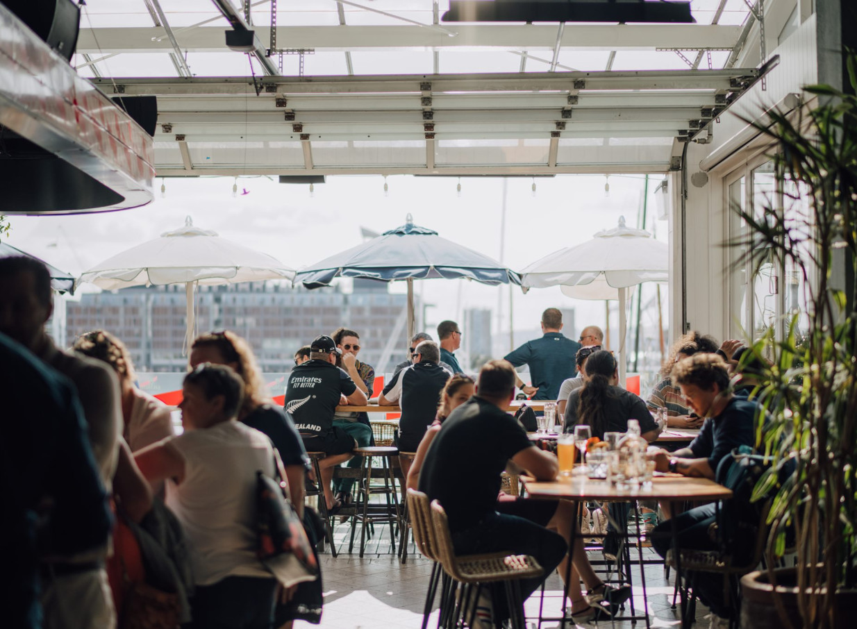 A lively, open-air rooftop bar and dining area at Dr Rudi's in Auckland's Viaduct Harbour, bustling with patrons overlooking the water.