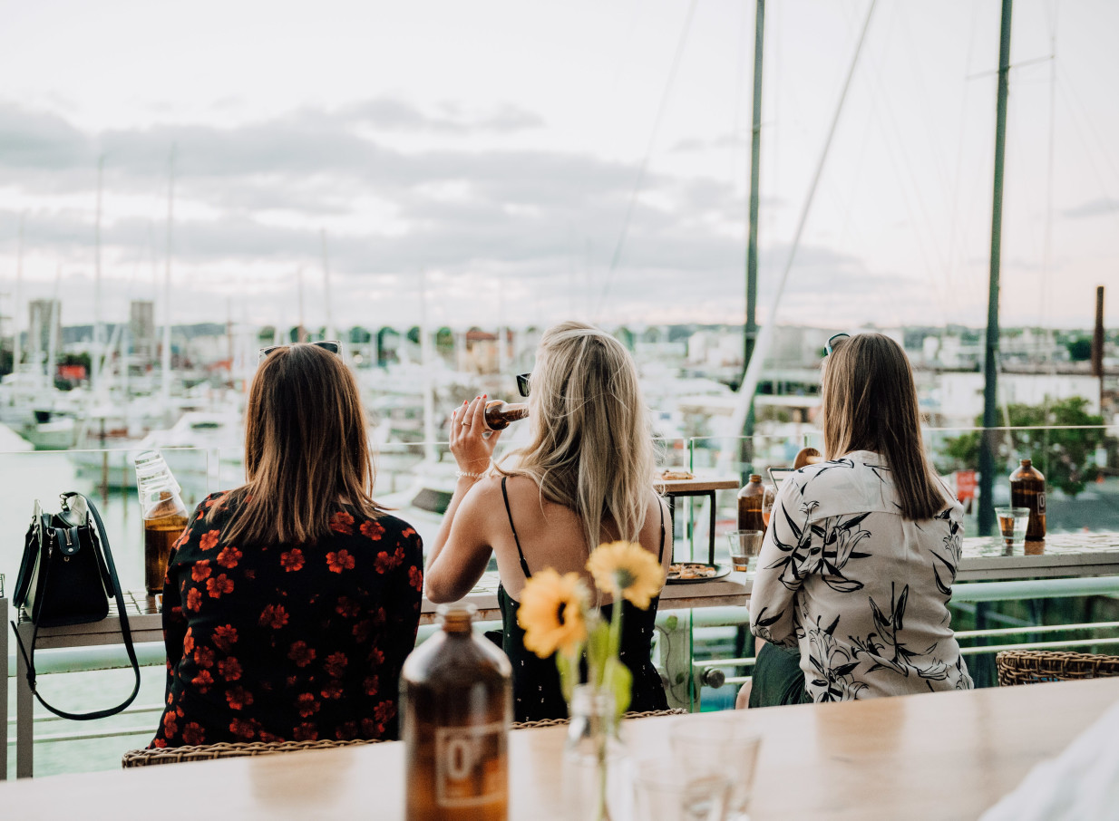 Three women enjoy drinks at the vibrant Dr Rudi's rooftop bar in Auckland, overlooking the Viaduct Harbour.
