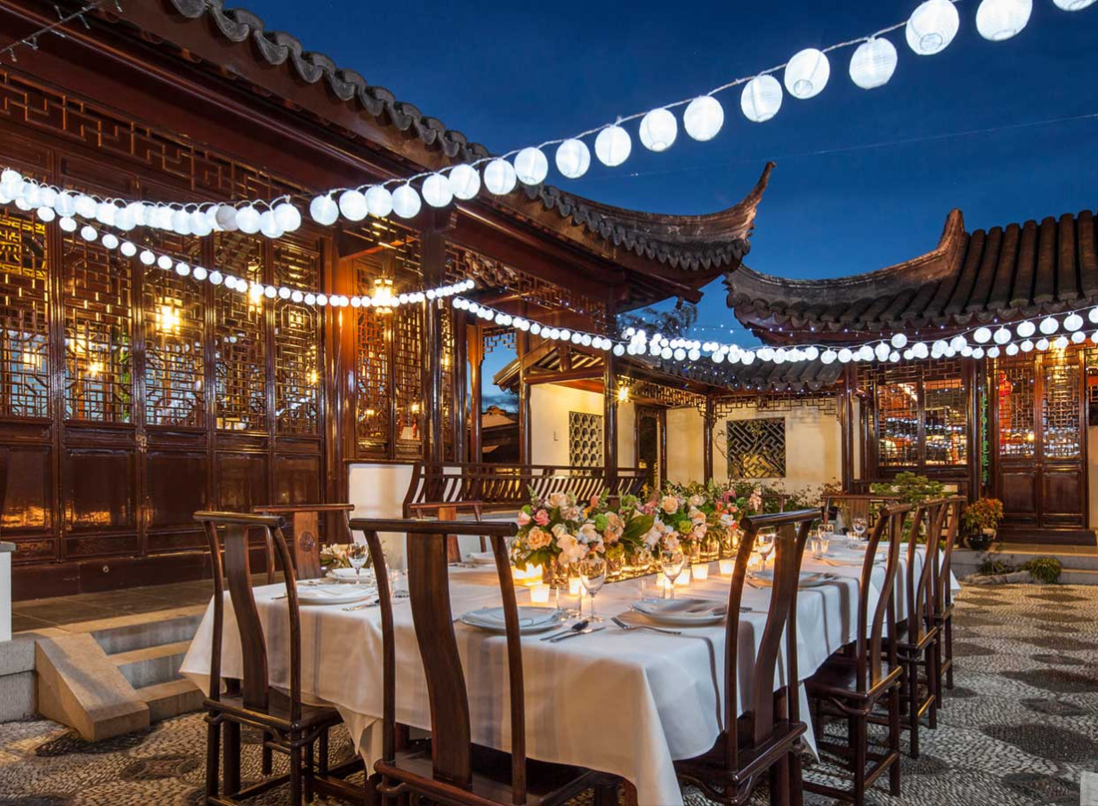 An elegant outdoor dining event is set within the traditional Chinese garden architecture of the Dunedin Chinese Garden, illuminated by festive lanterns.